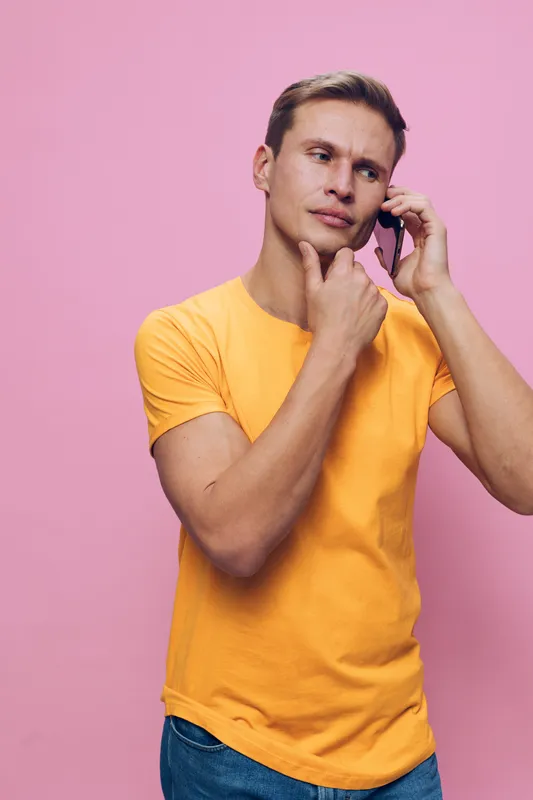 Thoughtful man with short hair wearing casual yellow t-shirt holding cellphone near ear looking away isolated on pink background studio portrait. People lifestyle concept