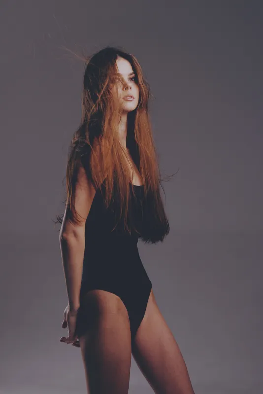 Portrait of a young woman in black bodysuit with long hair posing against gray background, elegant and thoughtful expression