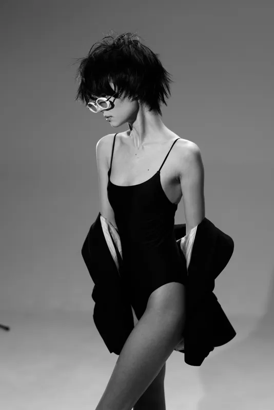 Black and white portrait of a slender woman wearing sunglasses and a black swimsuit, posing with a loose jacket against a neutral background.