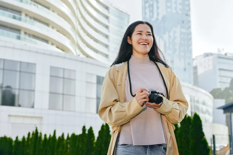 Smiling woman holding a camera in an urban outdoor setting with modern buildings, casual jacket and sweater, capturing candid moments in city life and everyday street photography against a bright