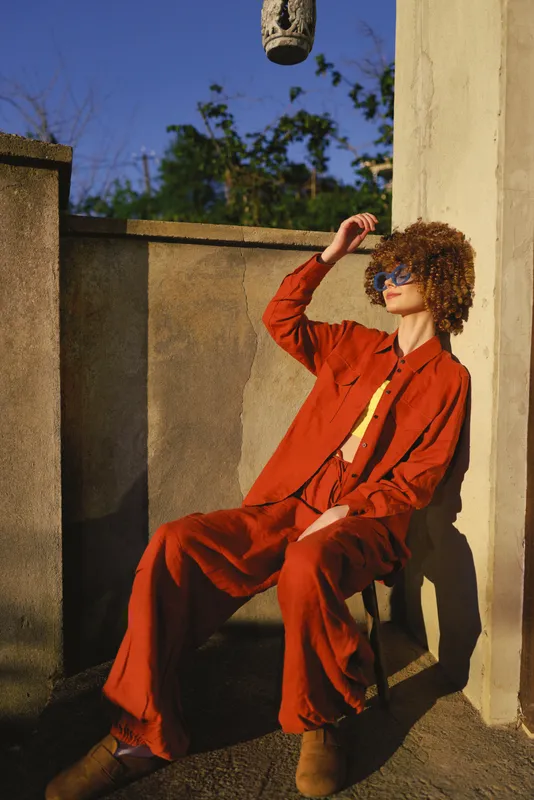 Young woman with curly hair sits on a sunlit concrete ledge wearing a bold orange jumpsuit, shading her eyes with one hand and looking relaxed