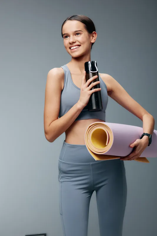Female fitness enthusiast in athletic wear holding a water bottle and rolled yoga mat, ready for a workout in a clean studio, highlighting health, balance and an active lifestyle.