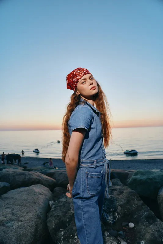 lifestyle woman wearing modern boho-western denim street style with red bandana on rocky beach during sunset, film color aesthetic, casual and relaxed vibe