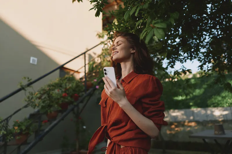 A young woman in a red dress smiles softly while looking up and holding a smartphone outdoors near lush greenery and sunlit stairs, conveying a casual, confident lifestyle moment