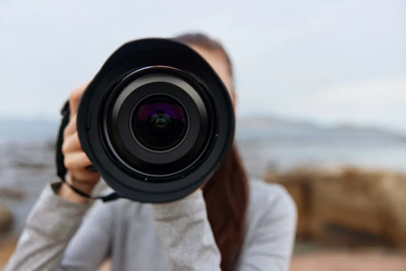 Woman holding a professional camera, focusing on photography skills outdoors with a blurred coastal background, expressing creativity and passion for capturing moments