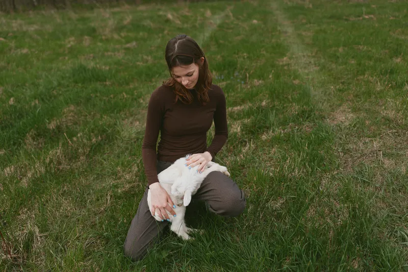 Young woman in brown sweater kneels on grass while gently holding and petting a white baby goat in a peaceful outdoor setting during daytime.