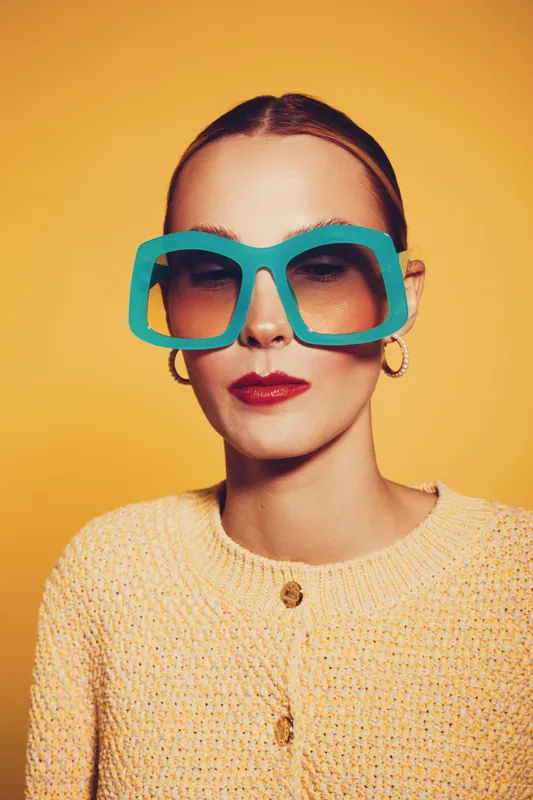Young woman with slicked hair wears oversized blue sunglasses and yellow sweater against orange background. Fashion portrait with bold accessories and bright colors.