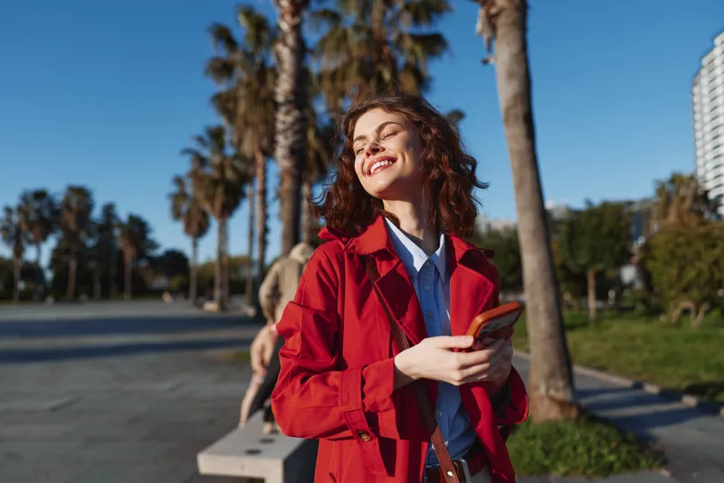 Happy smiling woman in a red jacket posing outdoors with a phone, bright sunlight, palm trees in the background, urban park vibe, casual fashion and positive mood.