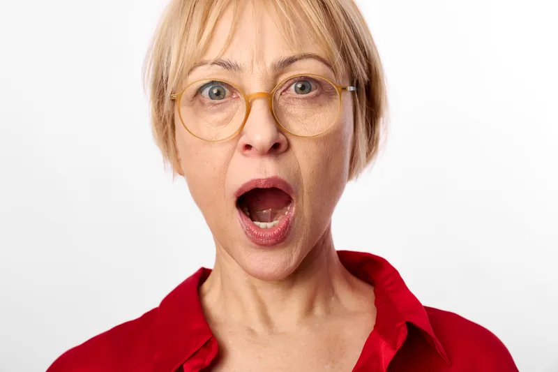 Surprised woman with blonde short hair and glasses, senior face in close up, red shirt against white background, expressing shock and emotion, studio portrait, copy space today