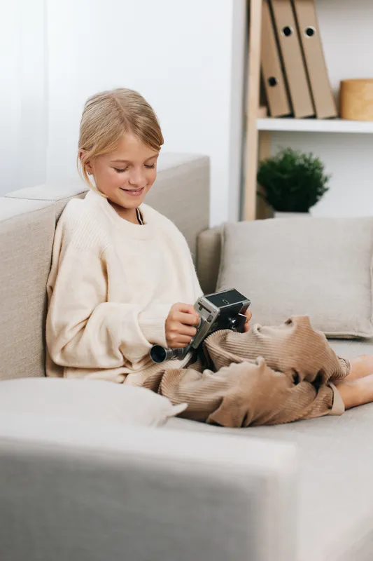 Young girl relaxing on a cozy sofa, focused on a handheld game controller, legs tucked, wearing a warm sweater, soft natural light creating a calm, playful moment for a cozy scene.