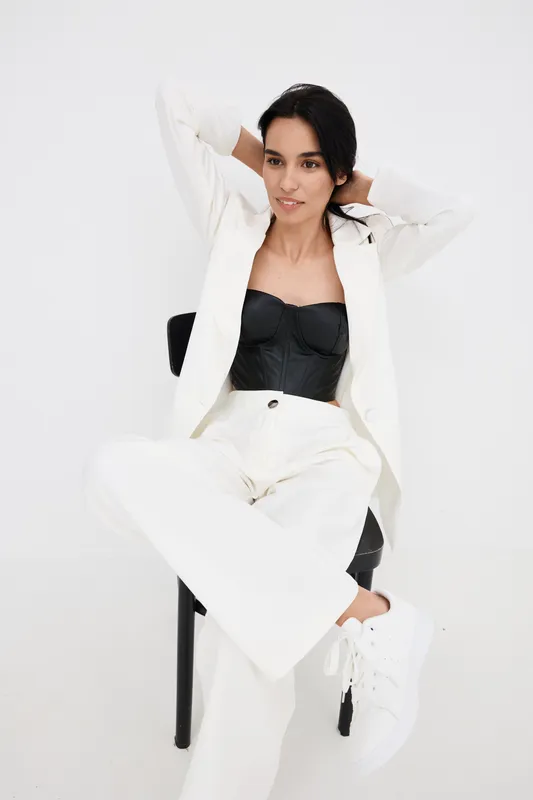 Young woman in white suit and black corset sitting relaxedly on chair against white background, showcasing modern fashion and casual elegance with sneakers.
