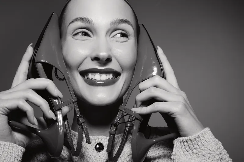 Smiling Caucasian woman with light skin holding two high heel shoes near her face in studio. Concept of happiness and fashion.
