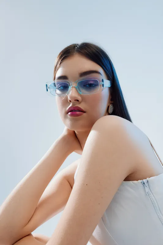 Young woman with long dark hair wearing transparent blue sunglasses and a white top poses against a light background. Concept of beauty, fashion, and modern style.