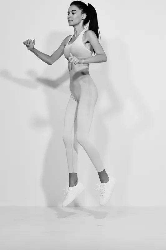 Sport fitness woman jumping in light workout clothes, focused expression, athletic pose on plain white background, demonstrating strength and motion.