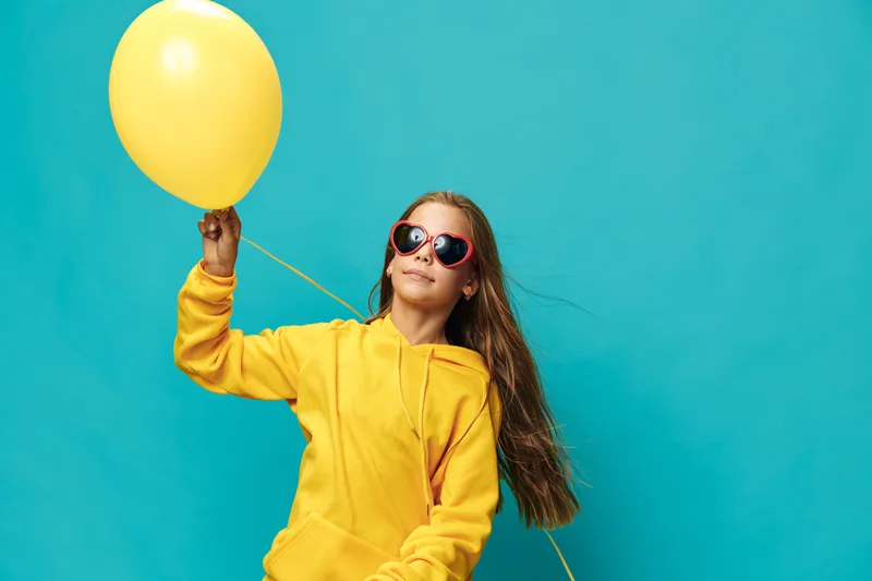 Girl in yellow hoodie holding yellow balloon with heart-shaped sunglasses on blue background. Child with long hair posing smiling for celebration or party concept.