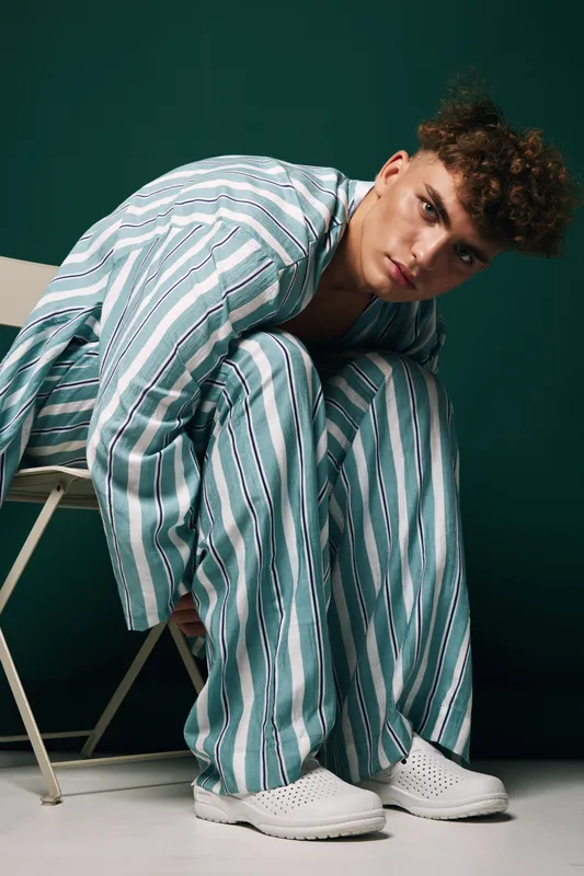 Young man in striped pajamas sitting on chair with serious expression against dark green background in studio. Casual fashion, relaxed pose and modern style.
