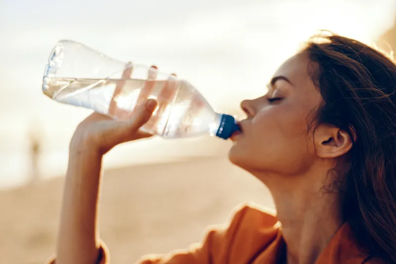 Woman drinking water bottle outdoors in summer sunlight, side profile close up, she takes a cool sip, hydrated smile, warm natural colors, soft blurred background bokeh