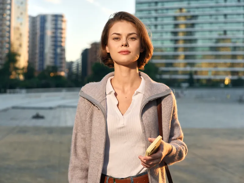 A confident professional woman stands outdoors in a modern urban setting. She smiles softly while holding a notebook, creating a calm, focused, and approachable work vibe.
