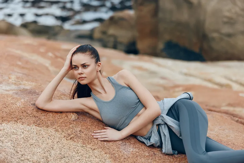 Woman in athletic wear lounging on a rock by the water, showcasing a calm expression in a natural outdoor setting, ideal for wellness and fitness themes