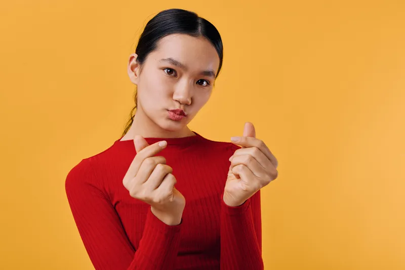 Young woman in red sweater making heart gesture with fingers against orange background, expressing love or affection, posing with serious facial expression and neat hair tied back.