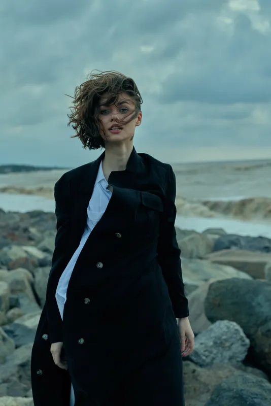 Windblown woman standing on rocky shoreline in a dark coat and white shirt, facing the sea under moody clouds, exuding bold style and coastal confidence.