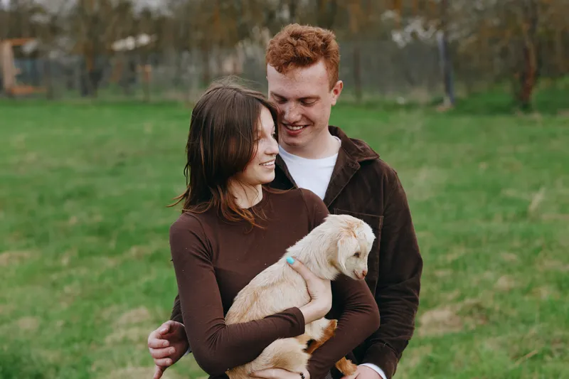 Young couple enjoys a tender moment outdoors as the woman holds a small goat in her arms while the man embraces her from behind in casual autumn clothing.