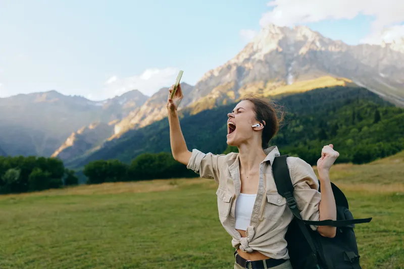 Joyful woman hiking in a mountainous landscape, backpacked and casual, shouting with excitement in an open field, clear sky and green valley behind her, lifestyle adventure scene