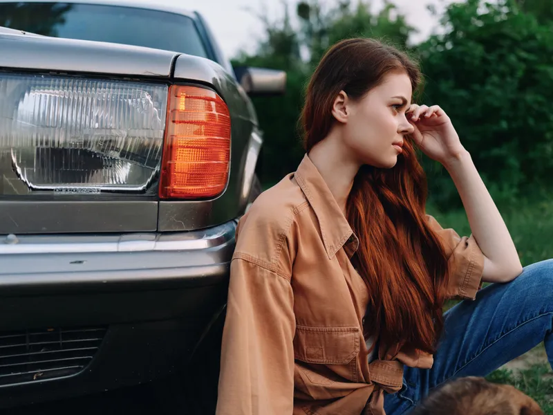 Young woman with long brown hair in casual attire sitting by a vintage car, expressing contemplation in a lush green setting.