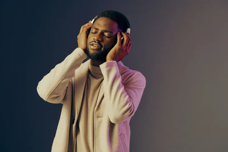 A young Black man enjoying music with headphones, showcasing a joyful expression, surrounded by a colorful gradient background, reflecting a modern lifestyle