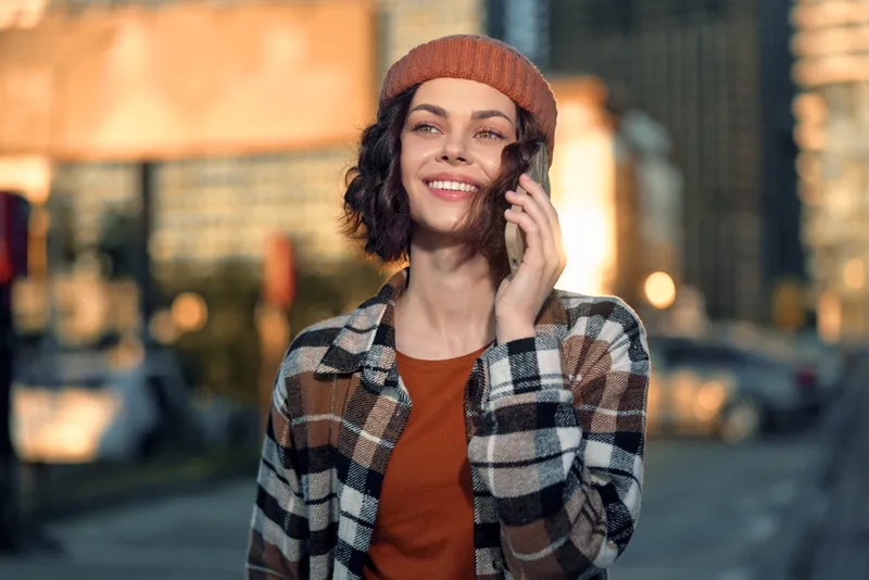 woman on phone with smile in urban street, candid lifestyle portrait at golden hour glow; beanie and plaid jacket convey authenticity, mindful living and emotional storytelling, soft warm light and