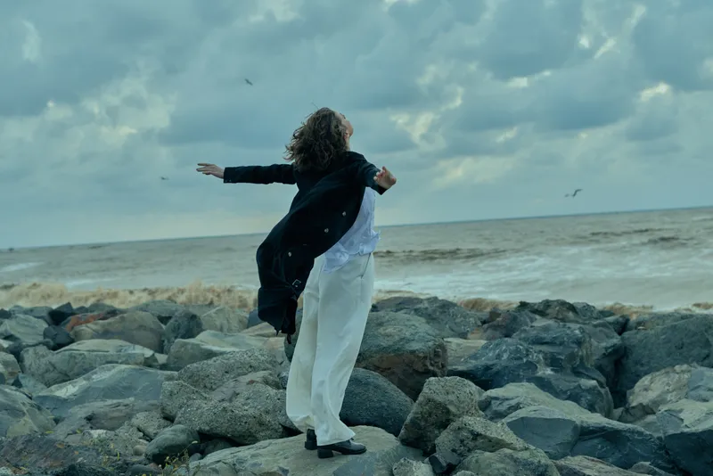 Dance, movement, shore, ocean, rocky, couple in motion on windy coast with waves and clouds, two figures embracing on stones, expressing freedom, balance, and connection during outdoor seaside moment