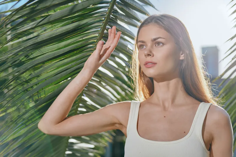 young woman posing near tropical palm leaves in warm sunlight wearing white sleeveless top with natural makeup and long straight hair, summer outdoor portrait