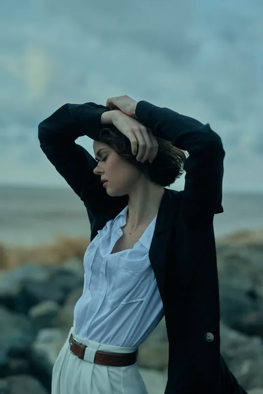 Woman standing outdoors by the coast with a blazer over a white shirt, arms raised in a confident pose, wind in hair, moody coastal setting, dramatic elegant fashion forward