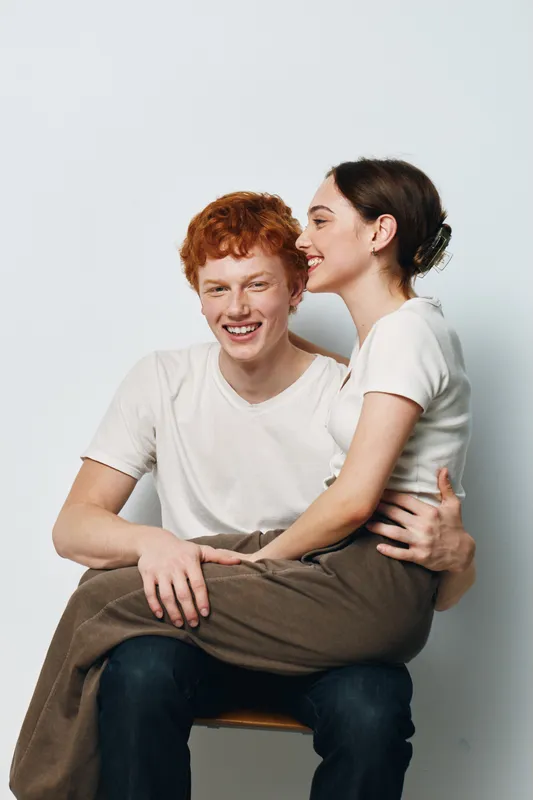 Young couple sitting close with smiling faces and casual clothes, happy affectionate relationship and joyful moment captured in simple white studio background.