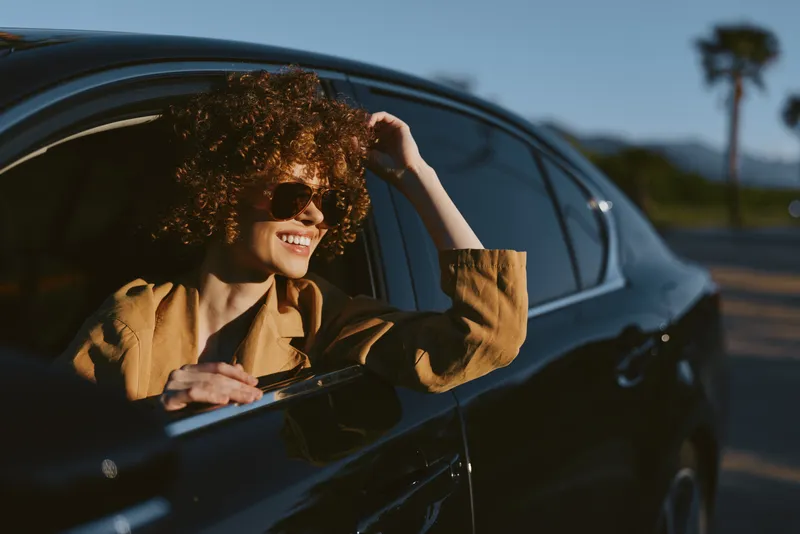 Smiling woman with curly hair leaning from a car window in warm retro tones, capturing a casual lifestyle and sunny mood with cinematic color.