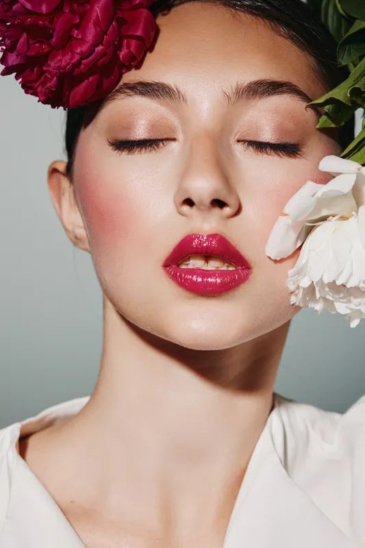 Close-up of a woman with pink lipstick and shimmering eye makeup posing with flowers on face. Creative beauty shot highlighting fashion makeup and floral style.