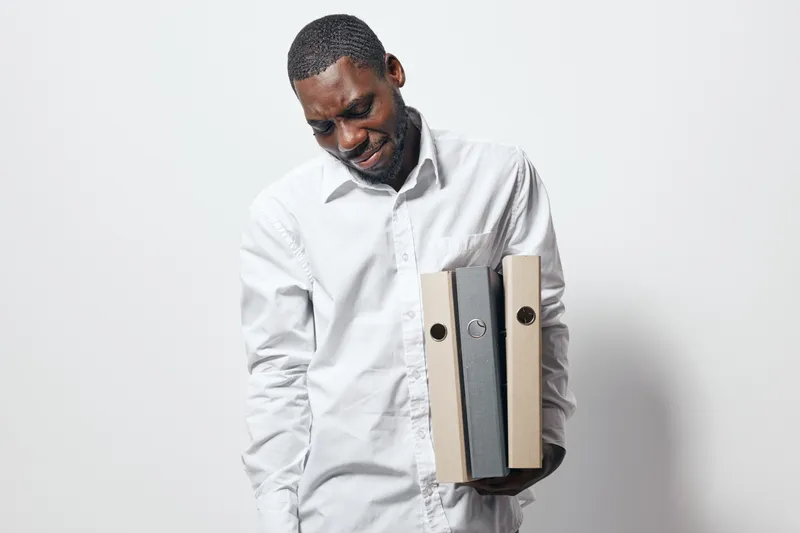 African American man with dark skin wearing white shirt holding cardboard folders for office work, documents and organization on white background with thoughtful expression.