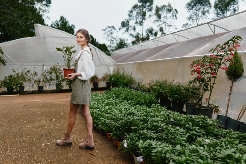 Young woman with a potted plant in a greenhouse, wearing a casual outfit, surrounded by greenery and flowers, showcasing a connection with nature