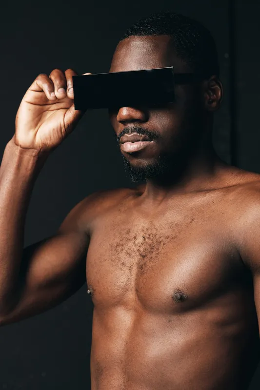 Creative conceptual visual style with African man shirtless holding black card over eyes in dark background. Artistic portrait with focus on skin texture and expression.