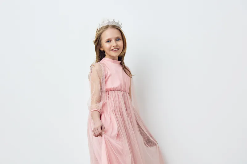 Charming young girl wearing a pink dress and a crown, joyfully smiling against a minimalistic white background, perfect for children s fashion concepts