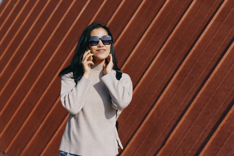 Woman in a cozy sweater talks on a phone, wearing sunglasses against a striped wooden wall, bright outdoor light, casual urban fashion, carefree mood and modern style