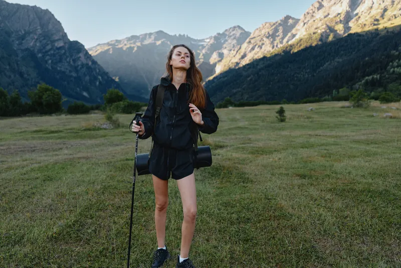 A young traveler stands in a scenic valley with towering mountains in the background, posing with a camera and backpack, enjoying sunlight and the fresh outdoor air.