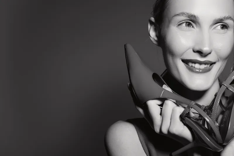 Portrait of a smiling white woman with fair skin holding high heel shoes near her face in a studio with dark background.