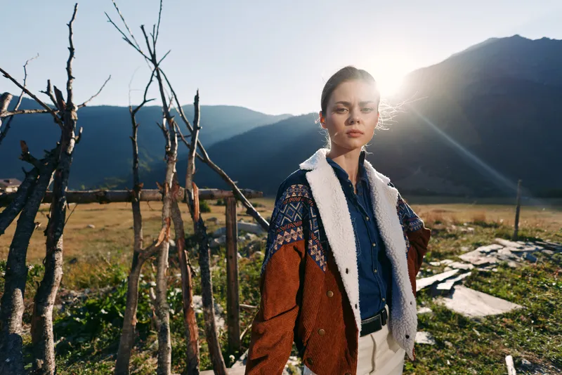 Woman portrait mountain countryside sunset jacket sweater rural fashion backlight. Young female model stands in sunlit valley by wooden fence wearing warm patterned knit jacket for outdoor style.