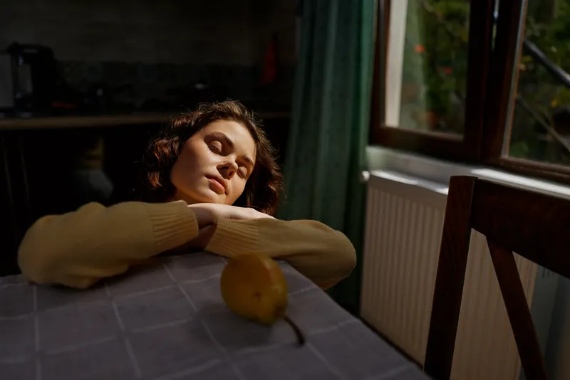 Relaxed woman with curly hair resting on a table in a softly lit room, yellow sweater, lemon on surface, warm mood, home environment, casual lifestyle, natural window light, intimate atmosphere,