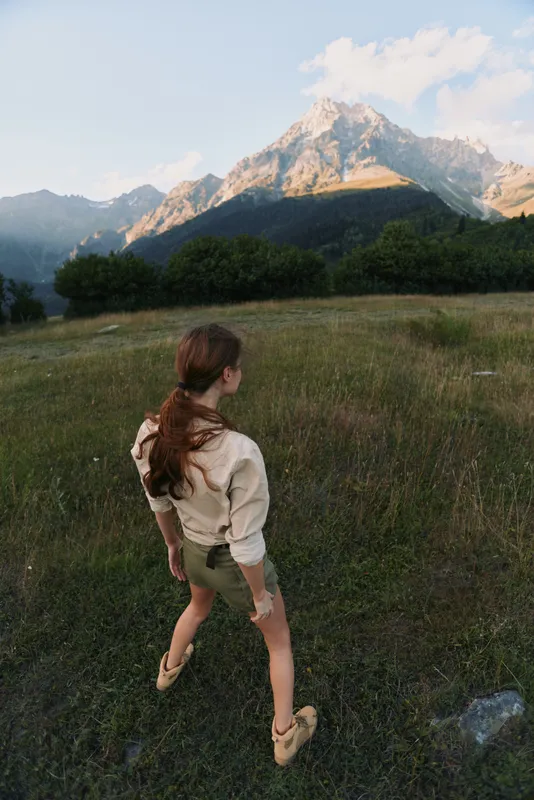 Young woman in a beige sweater and shorts walks across a sunlit meadow toward distant mountains, conveying curiosity, serenity and a sense of quiet outdoor adventure with gentle natural lighting.
