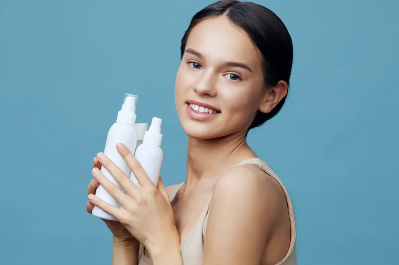 Young female model showcasing skincare products against a soft blue background, promoting healthy skin and self care with a bright smile