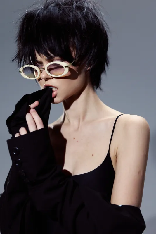 Stylish woman with short messy hair wearing black outfit and white sunglasses in minimalistic setting