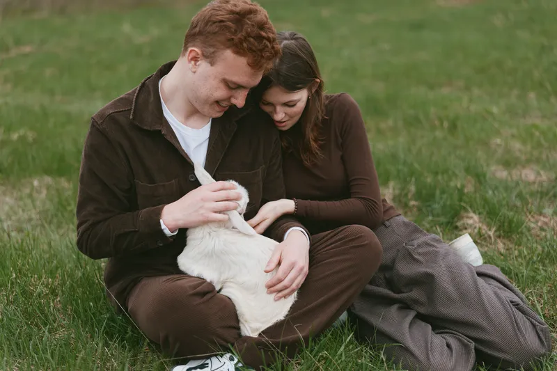 Affectionate couple outdoors with a gentle goat kid on green grass, sharing a warm moment and cuddling the playful animal. Natural light and casual setting convey lifestyle warmth and bonding.