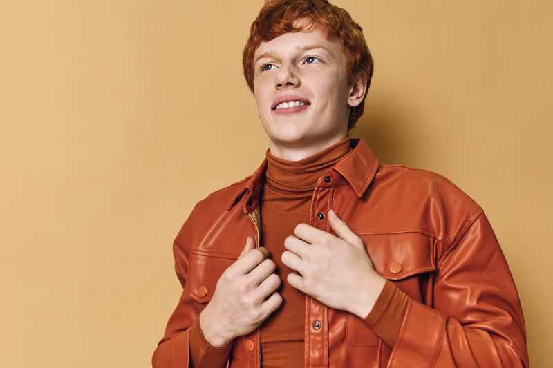 Young man with red hair wearing brown leather jacket and turtleneck sweater standing against beige background looking aside confidently.